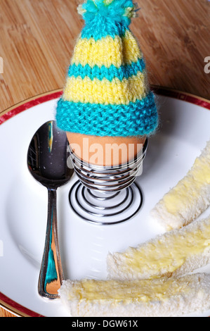 Gekochtes Ei in eine aufgerollte Metall Eierbecher garniert mit gemütlicher gestrickte Ei auf einem weißen Teller mit Weißbrot Soldaten. Stockfoto