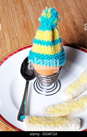 Gekochtes Ei in eine aufgerollte Metall Eierbecher garniert mit gemütlicher gestrickte Ei auf einem weißen Teller mit Weißbrot Soldaten. Stockfoto