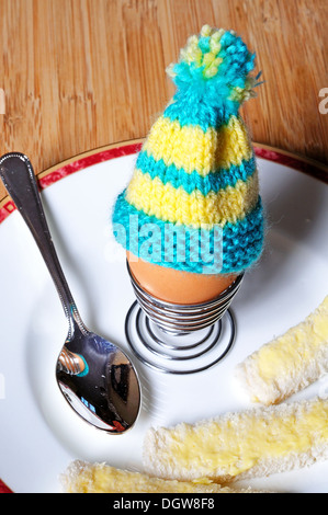 Gekochtes Ei in eine aufgerollte Metall Eierbecher garniert mit gemütlicher gestrickte Ei auf einem weißen Teller mit Weißbrot Soldaten. Stockfoto