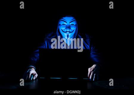 Junger Mann trug eine anonyme Maske und einer Kapuze sitzt vor einem laptop Stockfoto