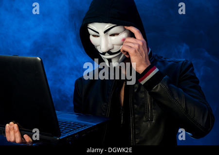 Junger Mann mit einer anonymen Maske mit einer Kapuze und eine schwarze Lederjacke, hält einen Laptop, eine nachdenkliche Geste Stockfoto