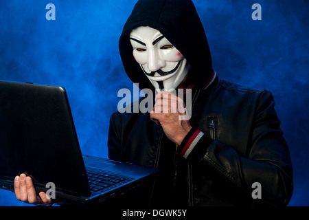 Junger Mann mit einer anonymen Maske mit einer Kapuze und eine schwarze Lederjacke, hält einen Laptop, eine in Anbetracht Geste Stockfoto