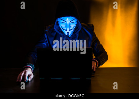 Junger Mann trug eine anonyme Maske und einer Kapuze sitzt vor einem laptop Stockfoto