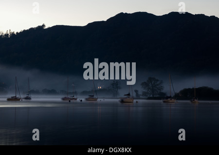 Eine magische spätabends Szene auf Windermere Lake mit fernen Ufer, Bäume und Yachten im Nebel versteckt, als letztes Licht verblasst. Stockfoto