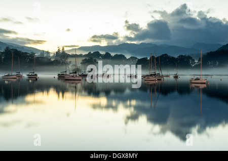 Einen späten Abend Szene auf Windermere See mit weit entfernten Bäumen und Yachten im Nebel verborgen und im Wasser spiegelt. Stockfoto