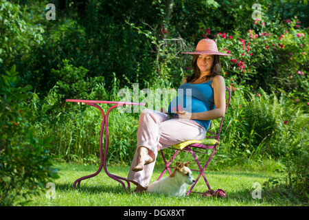 Schwangere Frau sitzt in einem Garten mit Hund Stockfoto