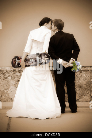 Küssen Hochzeitspaar von hinten gesehen, die Braut hält eine Skateboard, der Bräutigam trägt Handschellen und halten eine Braut Stockfoto