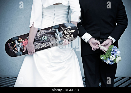Hochzeit paar von hinten gesehen, die Braut hält eine Skateboard, der Bräutigam trägt Handschellen und halten ein Brautstrauß Stockfoto