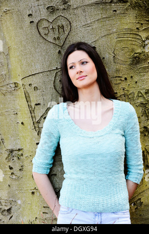 Junge Frau lehnt sich an einen Baum mit einem geschnitzten Herzen, Österreich Stockfoto