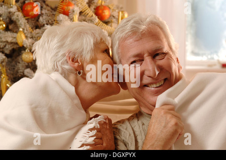 Älteres Paar in Liebe eingehüllt in einer Decke sitzt vor einem Weihnachtsbaum Stockfoto