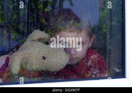 Junge Blick aus einem Fenster mit Wassertropfen, ein Stofftier im Arm halten Stockfoto