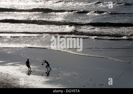 Silhouettenhafte Figuren von zwei jungen Männern, die Fußball spielen auf einem Strand vor dem Hintergrund des Meeres und hellem Sonnenlicht Stockfoto
