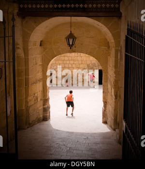 Zwei Jungen spielen Fußball (Fußball) in einem sonnigen Innenhof in Vittoriosa (Birgu), Malta.  Das Malteser Kreuz ziert den Torbogen oben. Stockfoto