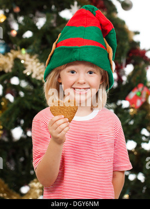 Ein netter lächelnder junge verkleidet als Weihnachtsmann Helfer Lebkuchen vor einem Weihnachtsbaum hält. Stockfoto