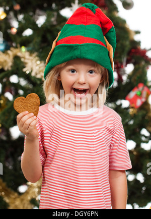 Ein niedliches begeistert Junge gekleidet wie santa's Helper ist Holding Lebkuchen cookie Vor einem Weihnachtsbaum. Stockfoto