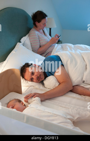 Neue Eltern im Bett, Papa tröstlich Baby in der Krippe und Mama mit Handy Stockfoto