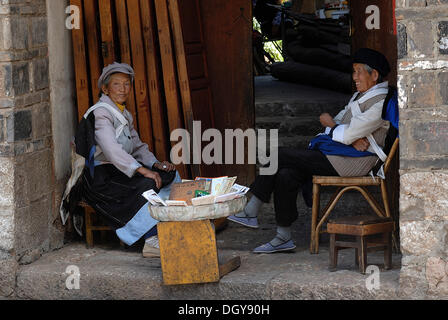 Zwei Frauen in der Tracht der Naxi-Minderheit sitzen mit einem kleinen Verkaufsstand in einem Hauseingang, Lijiang, Yunnan Stockfoto