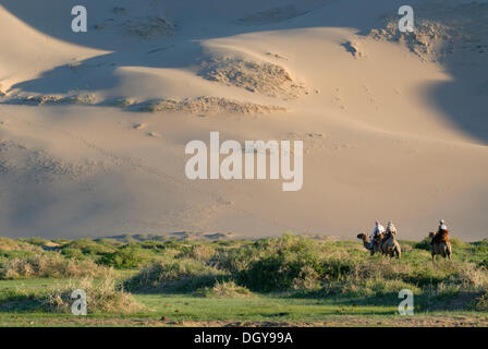 Touristen auf Kamel reiten durch ein sattes Grün grass Landschaft in Richtung der großen Khorgoryn Els Sanddünen in der Wüste Gobi Stockfoto