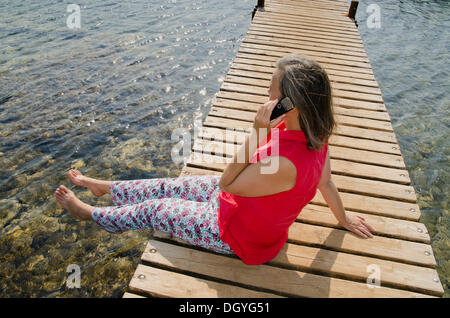 Frau sitzt auf einem Dock am Wasser mit einem Handy, Tisno, Murter Insel, Adria, Dalmatien, Kroatien, Europa Stockfoto