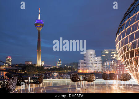 Terrasse eines Restaurants, Gehry-Bauten in der Abenddämmerung auf der Rückseite, Neuer Zollhof Gebäudekomplex und der Rheinturm-Turm Stockfoto