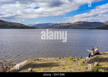 Ein älteres paar männliche und weibliche sitzen an der Seite von Loch Katrine, ist sie auf ein Feature zeigt, während das Männchen eine Kamera verwendet Stockfoto