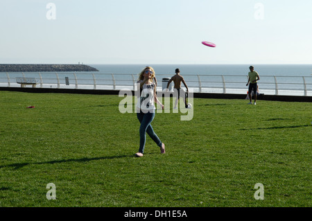 Junge Frau spielen Frisbee in einem Park direkt am Meer in Wales, zwei junge Männer sind einen Ball im Hintergrund Stockfoto