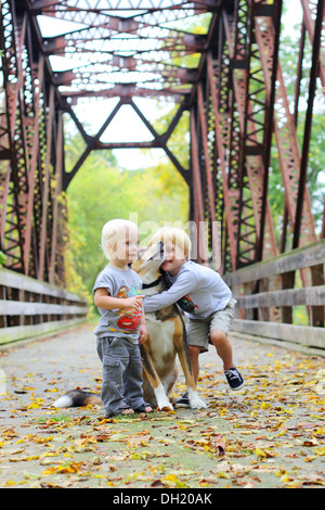 zwei Kinder, einen jungen und seinen kleinen Bruder als draußen auf einer Brücke in den Wald, liebevoll umarmt ihre Schäferhund Stockfoto