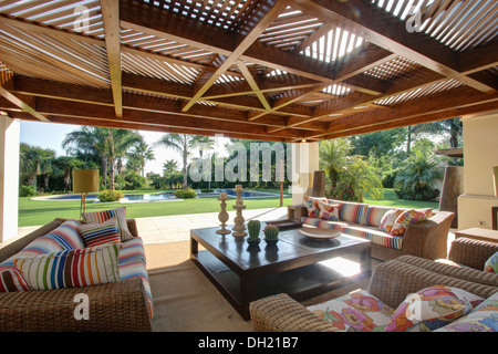 Gestreiftes Kissen auf komfortablen Wicker Sofas neben Couchtisch auf Veranda mit Balkendecke und Blick auf pool Stockfoto