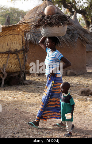 Frau mit Sorghum auf dem Kopf mit ihrem Kind in einem Dorf, Burkina Faso Stockfoto