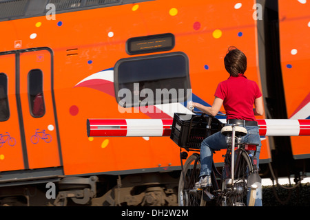 Niederlande, Vogelenzang, Train Bahnübergang. Frau auf Fahrrad wartet Barriere. Royal Kings Orange Zug vorbei. Stockfoto