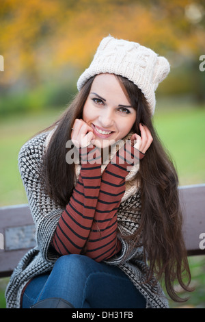 Junge Frau in einem Park im Herbst sitzen auf einer Bank einen wolligen Hut selbstbewusst in die Kamera Lächeln Stockfoto