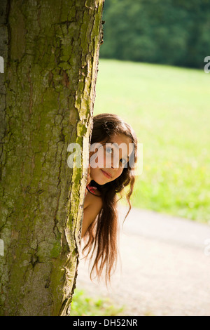 Lächelndes Mädchen schaut hinter einem Baum Stockfoto