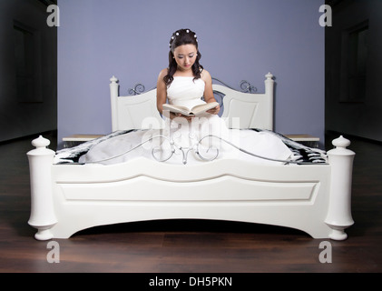 Mädchen in einem langen Kleid auf einem weißen Bett liest das Buch Stockfoto