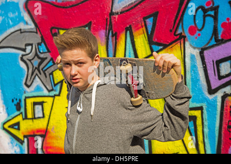 Junge, 13 Jahre, hält eine Skateboard vor Graffiti bewachsenen Wand, Deutschland Stockfoto