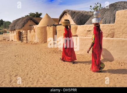 Zwei indische Frauen tragen rote Saris tragen von Wasser gefüllten Krügen auf dem Kopf durch ein Dorf, Thar-Wüste, Rajasthan, Indien Stockfoto