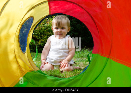 Neun Monate altes Baby Mädchen im Garten mit Blick durch große bunte Tuch Spielzeug Rohr. August. VEREINIGTES KÖNIGREICH. Stockfoto