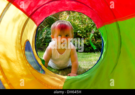 Neun Monate altes Baby Mädchen im Garten mit Blick durch große bunte Tuch Spielzeug Rohr. August. VEREINIGTES KÖNIGREICH. Stockfoto