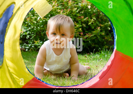 Neun Monate altes Baby Mädchen im Garten mit Blick durch große bunte Tuch Spielzeug Rohr. August. VEREINIGTES KÖNIGREICH. Stockfoto