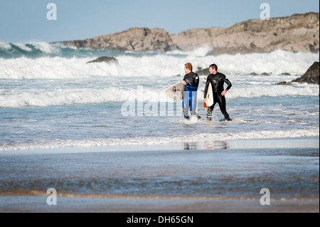 Zwei Surfer waten zu Fuß entlang der Küstenlinie von Fistral Strand in Newquay. Stockfoto