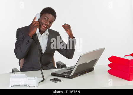 Junge dunkelhäutige Mann sitzt an einem Laptop und ein Handy beim suchen glücklich halten Stockfoto