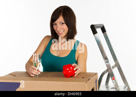 Junge Frau mit einem Umzugskarton, ein Sparschwein und einem Glas Champagner in der hand Stockfoto