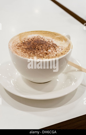 Eine Tasse Cappuccino mit bestreut Schokopulver auf Milchschaum auf einem Tisch mit einem mitreißenden Stock in die Untertasse. Stockfoto
