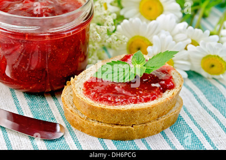 Zwei Stück Brot mit Erdbeermarmelade, gestreift ein Glas Marmelade, ein Messer und einen Blumenstrauß der Kamille auf einer grünen Serviette Stockfoto
