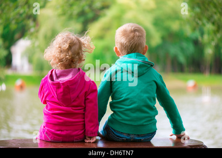 Kleine Schwester und Bruder sitzt auf der Bank am Teich. Sicht nach hinten. Stockfoto