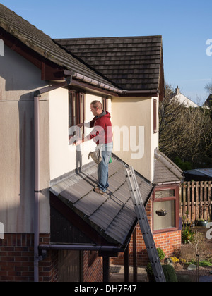 Eine männlichen Fenster auf Veranda Dach Reinigung Reiniger steht im ersten Stock nach oben Fenster eines Hauses mit einem Rakel. UK-Großbritannien Stockfoto