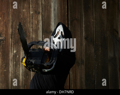 Mann in beängstigend "Schrei" Maske hält Kettensäge. Maske der Sensenmann. Weißen Geistermaske Karneval und schwarze Kapuze. Scary Movie Film. Stockfoto