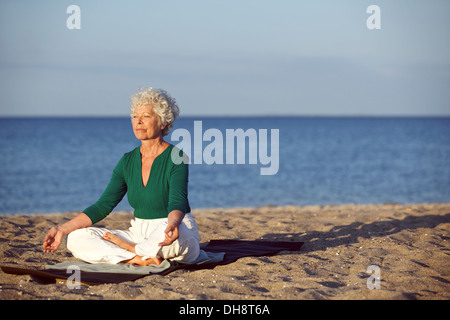 Bild der alten Frau am Strand sitzen auf Übung Matte Yoga zu praktizieren. Senior kaukasischen Frau meditieren im Lotus pose Stockfoto