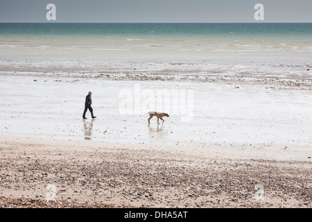 Mann zu Fuß seinen Labrador Hund bei Ebbe entlang einem Schindel gesäumten Sandstrand mit türkisblauem Meer im Hintergrund. Stockfoto