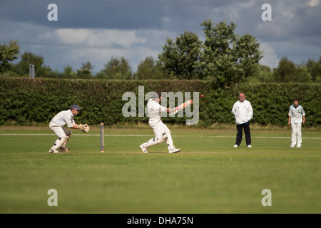 Dorf-Cricket. Die Schlagmann zieht den Ball Platz das Wicket mit dem Wicketkeeper auf der Lauer während ein Feldspieler und ein Stockfoto