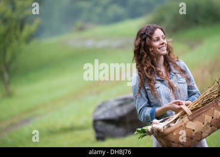 Eine junge Frau trägt einen Korb mit frisch geernteten Knoblauch und Gemüse. Stockfoto